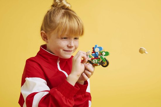 Blond meisje in rode jas houdt een LEGO Yoshi-Crosser vast, kijkt geconcentreerd.
