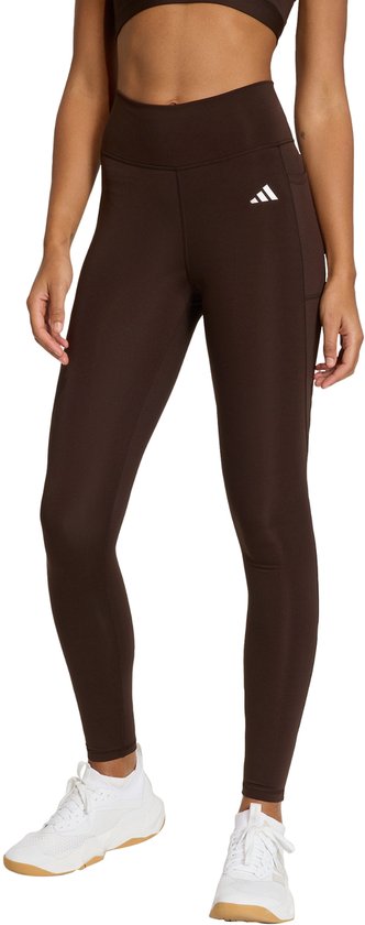 Vrouw in donkerbruine legging met wit Adidas logo, staand, witte sneakers.