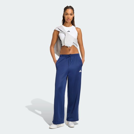 Model in een donkerblauwe trainingsbroek met witte strepen, Adidas logo en witte sneakers, staand met handen in de zakken.