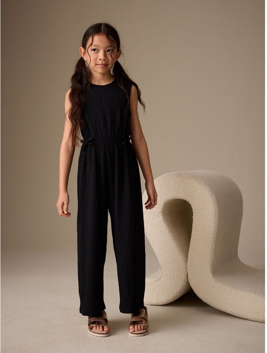 Zwart kreukjumpsuit met strikjes aan de zijkant, gedragen door een meisje met staartjes.