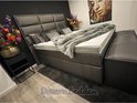 Grijze boxspring met hoog gestoffeerd hoofdbord, witte matrassen, grijze deken, sierkussens, bloemen en een nachtkastje.