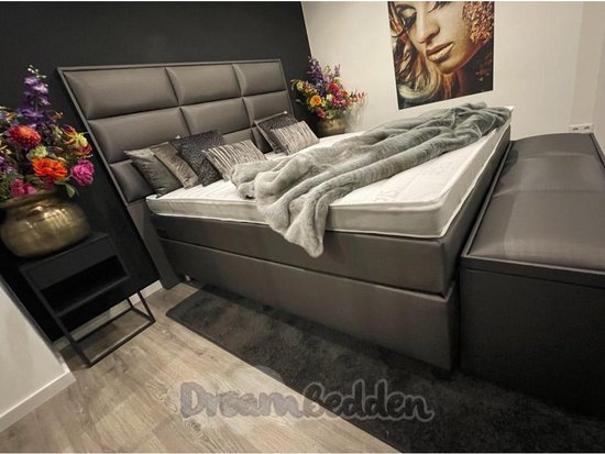 Grijze boxspring met hoog gestoffeerd hoofdbord, witte matrassen, grijze deken, sierkussens, bloemen en een nachtkastje.