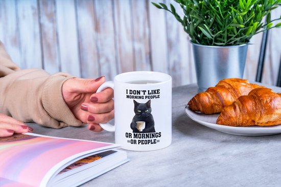 Witte mok met tekst en kat wordt vastgehouden door een persoon naast croissants.