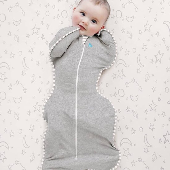 Baby met grote blauwe ogen ligt in grijze Swaddle UP slaapzak met streepjesrand op achtergrond met sterren en manen.