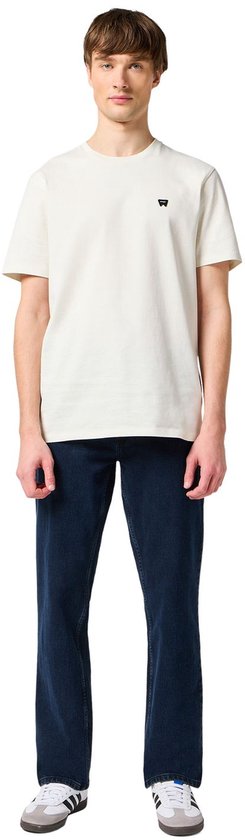 Man in een crème T-shirt met klein zwart logo en een donkerblauwe spijkerbroek.