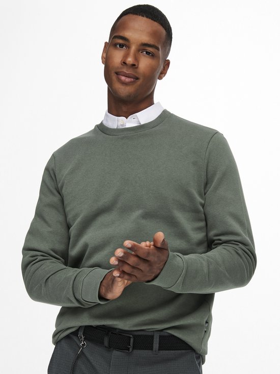 Man kijkt naar de zijkant met handen tegen elkaar. Draagt een groene sweater met wit overhemd eronder en grijze broek.