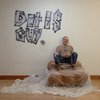 Man met tatoeages en spijkerbroek zit in bruin leer omwikkelde stoel, met "Dit Is Guy" graffiti op de muur.