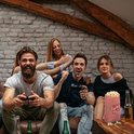 Vier vrienden spelen een videogame en eten popcorn. Een zakje met rode en witte strepen staat op tafel.