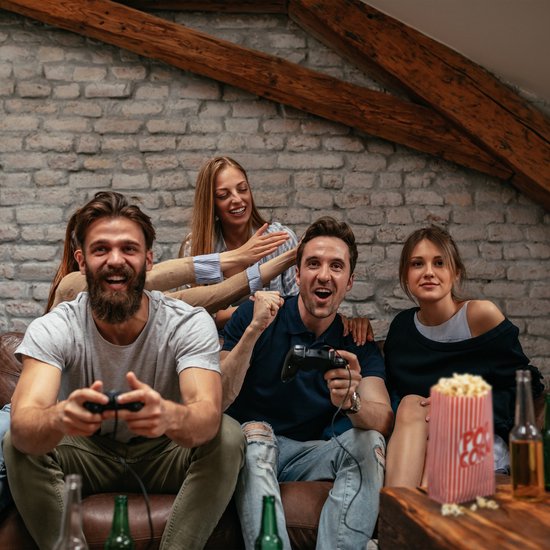 Vier vrienden spelen een videogame en eten popcorn. Een zakje met rode en witte strepen staat op tafel.