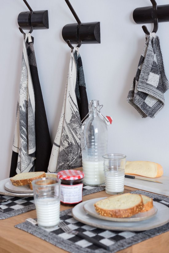Keukendoeken met verschillende patronen, waaronder een met blokjes, hangen aan de muur naast een ontbijttafel met brood, melk en jam.