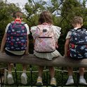 Drie kinderen zitten op een houten balk met hun rug naar de camera. Ze dragen rugzakken: een met raceauto's, een met fruitprint en een met ruimtevaartthema.