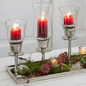 Drie rode kaarsen in geribbelde glazen houders op een zilverkleurig dienblad met dennenappels, mos en sneeuw.