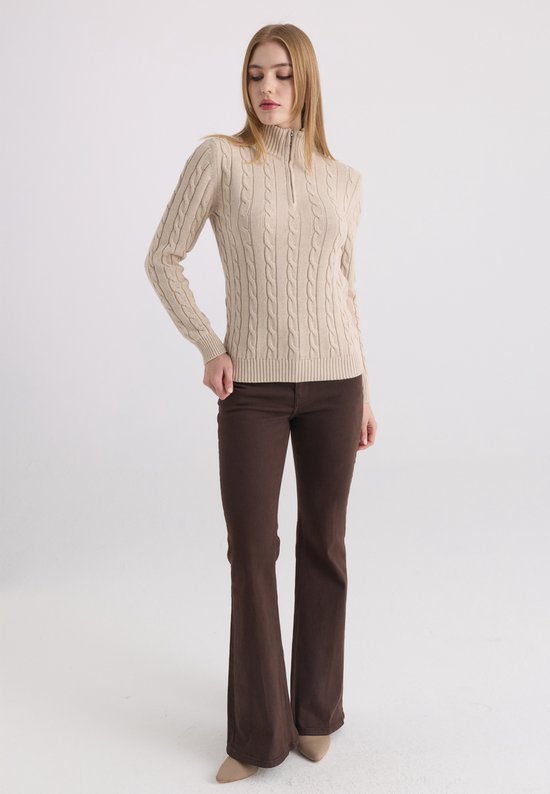 Model staat in driekwart profiel met een beige kabelgebreide trui met ritskraag en bruine broek.
