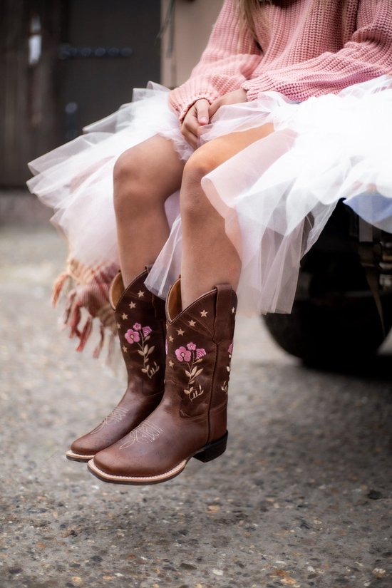 Kind draagt bruine leren cowboylaarzen met roze bloemen en sterren, gecombineerd met een witte tule rok en roze trui.