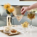 Een tafel met verschillende gouden, kristallen glazen, gevuld met drankjes en decoraties. Een hand voegt een takje toe aan een glas.