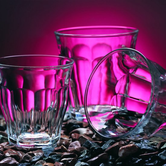 Trois verres Duralex Picardie transparents avec cannelures verticales, deux remplis de liquides colorés, sur un lit de pierres.