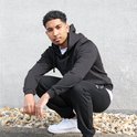 Jonge man in zwart hoodie en joggingbroek, gehurkt voor een grijze muur.