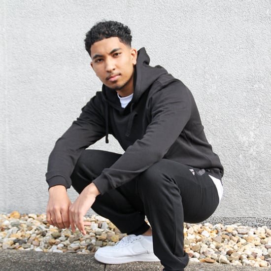 Jonge man in zwart hoodie en joggingbroek, gehurkt voor een grijze muur.