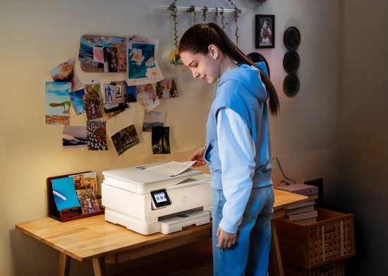 Jonge vrouw in blauwe hoodie plaatst document in de scanner van een witte HP Envy Photo printer op houten bureau.