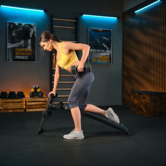 Vrouw in geel sport-top en grijze legging doet een halter-oefening op de schuine trainingsbank.