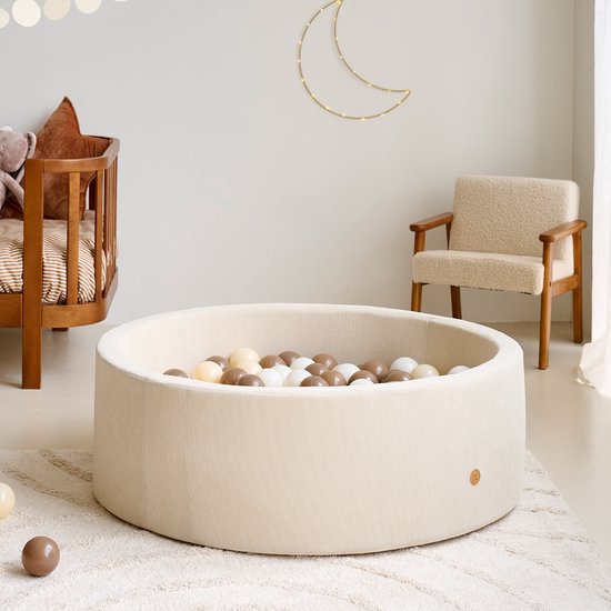 Een beige, ronde corduroy ballenbak vol met witte, beige en bruine ballen, in een kinderkamer.