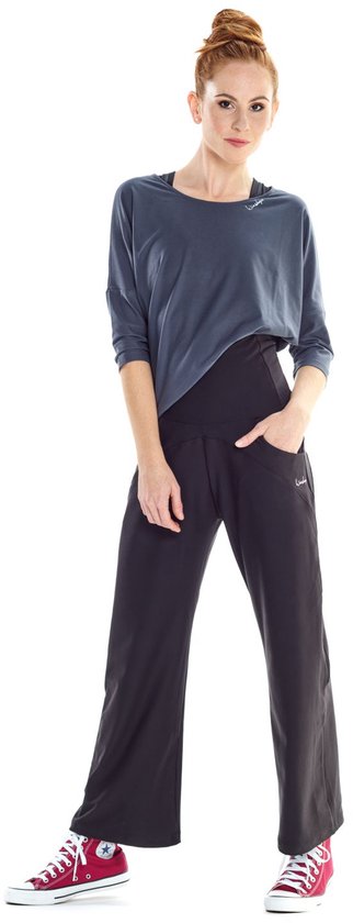 Model met rood haar in antracietkleurig shirt met driekwart mouwen en wijde zwarte broek, met rode sneakers.