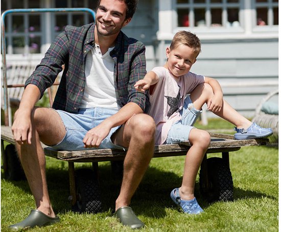 Man en jongen zitten op een kar in de tuin. Ze dragen tuinklompen in het blauw en groen.