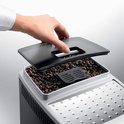 Een hand opent het deksel van het bonenreservoir van de zilveren De'Longhi Magnifica S espressomachine, gevuld met koffiebonen.