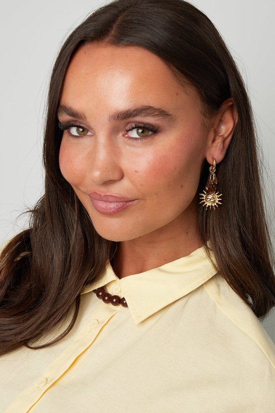 Model draagt gouden oorbellen met zonnevormige hangers en donkere kralen, aan een geel hemdje.