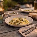 Fettuccine met courgette, Parmezaanse kaas, kruiden en olijven op een beige bord, met een brandende lantaarn ernaast.