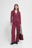 Vrouw met lang golvend bruin haar, draagt een bordeauxrode blazer, broek, bruine top, zwarte schoenen en tas.