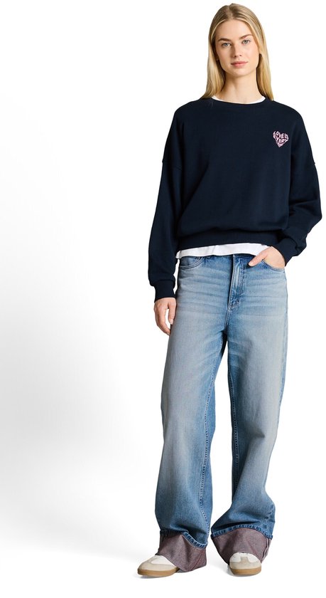 Model in een donkerblauwe sweater met een roze hartje en een wijde, lichtblauwe jeans met omgeslagen zoom.