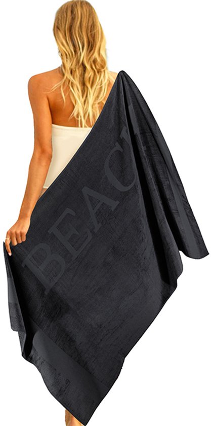 Vrouw met blonde haren, in een witte badpak, draagt een zwarte handdoek met 'BEACH' erop.