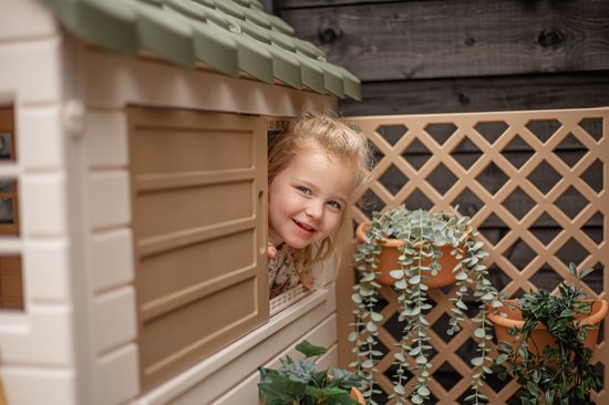 Meisje met gebloemd jurkje kijkt lachend door crème speelhuisje met beige luikje.