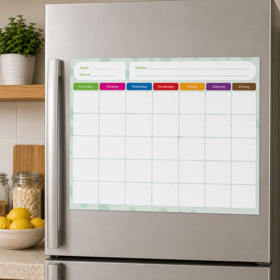 Een lichtgroene whiteboard weekplanner met een raster van 5x7 vakken hangt op een zilveren koelkast. De dagen van de week zijn gekleurd.