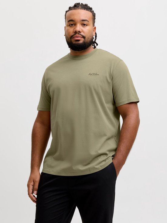 Man draagt olijfgroen T-shirt met 'Jack & Jones ARCHIVE SELECTION' logo en zwarte broek. Handen in zakken.