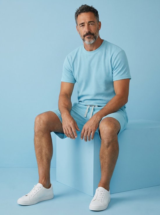 Man met grijze baard zit op een lichtblauwe kubus, draagt turquoise T-shirt en short met wafelpatroon en witte sneakers.