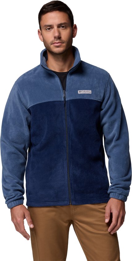 Een man draagt een blauwe fleece jas met een donkerblauwe borst en kraag. De jas heeft een rits en een Columbia-logo op de borst.