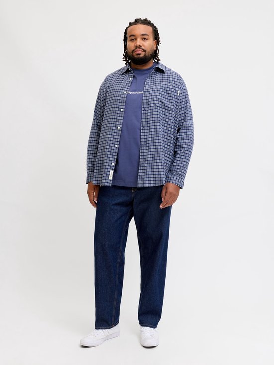 Man met dreadlocks draagt een blauw geruite blouse over een blauw T-shirt en donkere jeans.