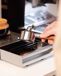Een hand houdt een zilverkleurige portafilter met houten handvat boven een uitklaplade van geborsteld staal.
