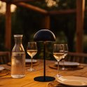 Lampe de table LED noire sans fil sur une table en bois à l'extérieur, avec une carafe d'eau et des verres à vin.