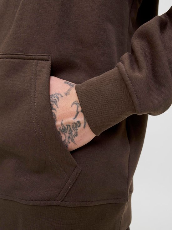 Hand met tatoeages steekt uit de steekzak van een bruine hoodie.