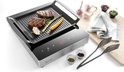 Inductie grillplaat met digitale display, waarop twee steaks, asperges, wortel en knoflook liggen. Naast de grill staan peper- en zoutvaatjes, tang en kruiden.