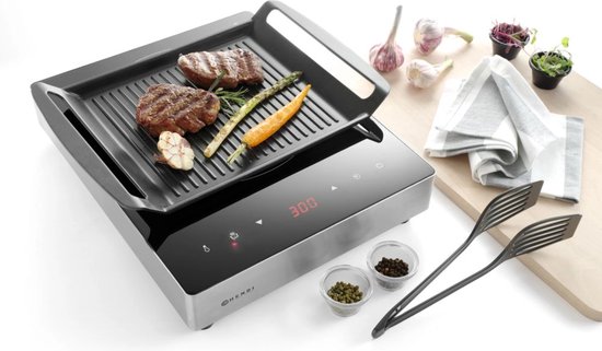 Inductie grillplaat met digitale display, waarop twee steaks, asperges, wortel en knoflook liggen. Naast de grill staan peper- en zoutvaatjes, tang en kruiden.