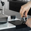 Handen die een portafilter tappen op een zwarte tampermat naast een espressomachine.