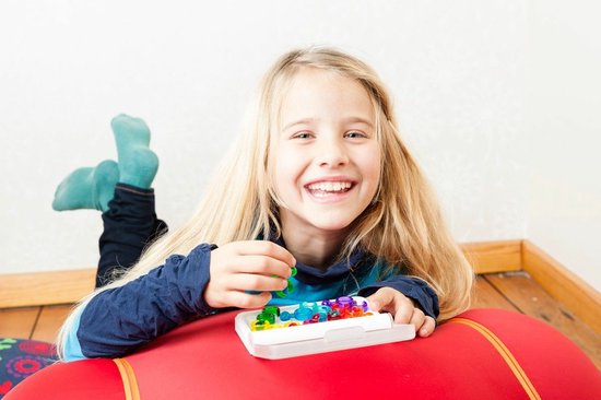 Meisje met lang blond haar, lachend, liggend op een rode bal, spelend met gekleurde spelonderdelen.