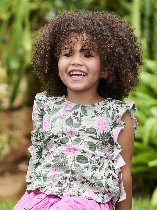 Vrolijk meisje met krullend haar draagt een lichtgroene blouse met bloemenprint, roze ruches en een roze rok.