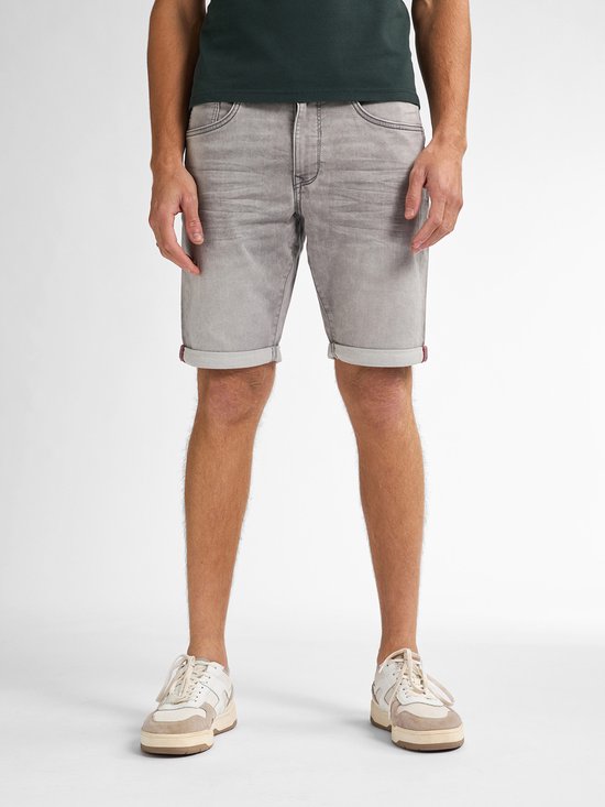 Man van voren, draagt een donkergroene polo en een grijze denim short met opgerolde zomen en rode accenten aan de binnenkant.