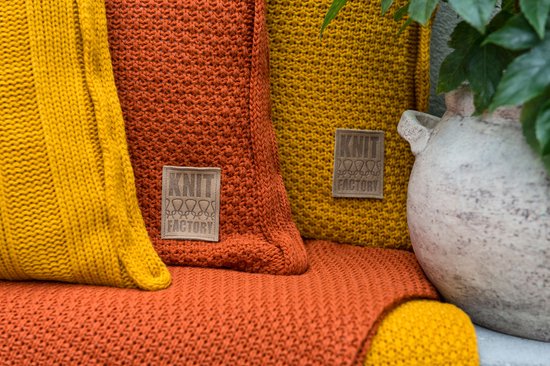 Deux plaids tricotés orange et un moutarde avec des étiquettes marron, à côté d'une plante verte.