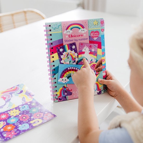 Une fille pointe du doigt un livre "Sticker by number" Licorne qu'elle tient. Le livre présente un arc-en-ciel, des nuages et des licornes sur la couverture avant.